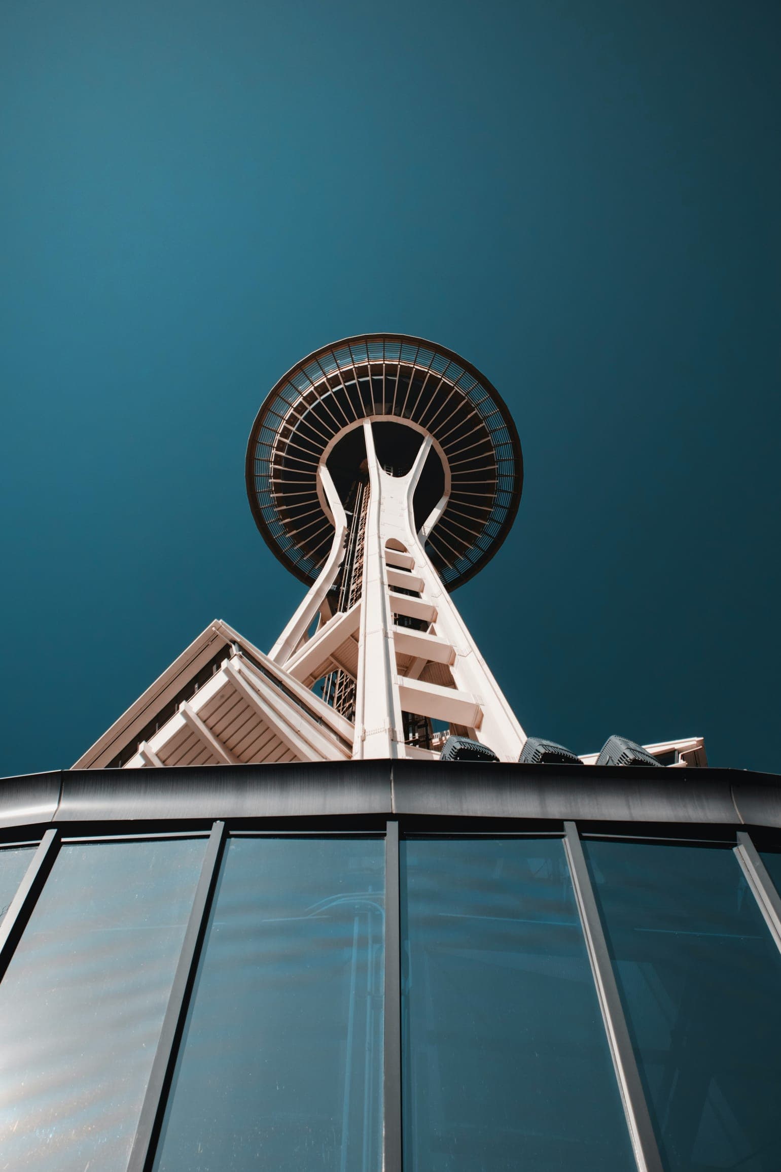 Space Needle tower seen from below against a deep teal sky, rising behind a glass-walled building.