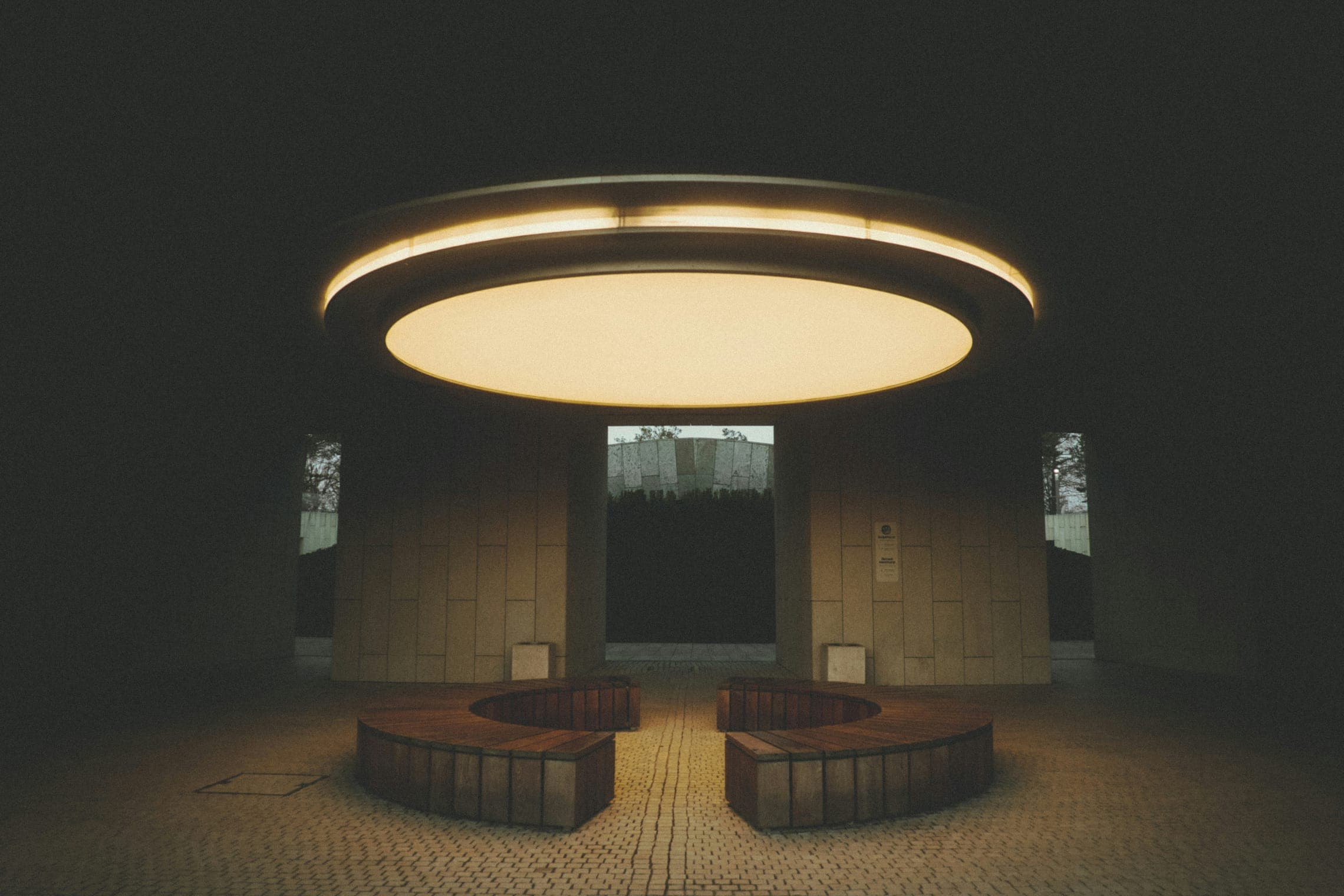 Round pavilion at night with a glowing ring-shaped ceiling light above two curved wooden benches.