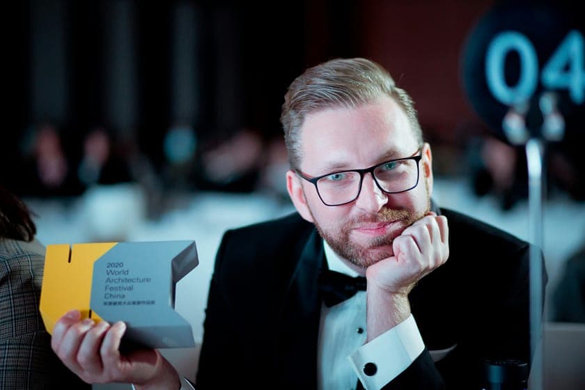 Smiling man in a tuxedo and glasses holding a yellow and grey architecture award.