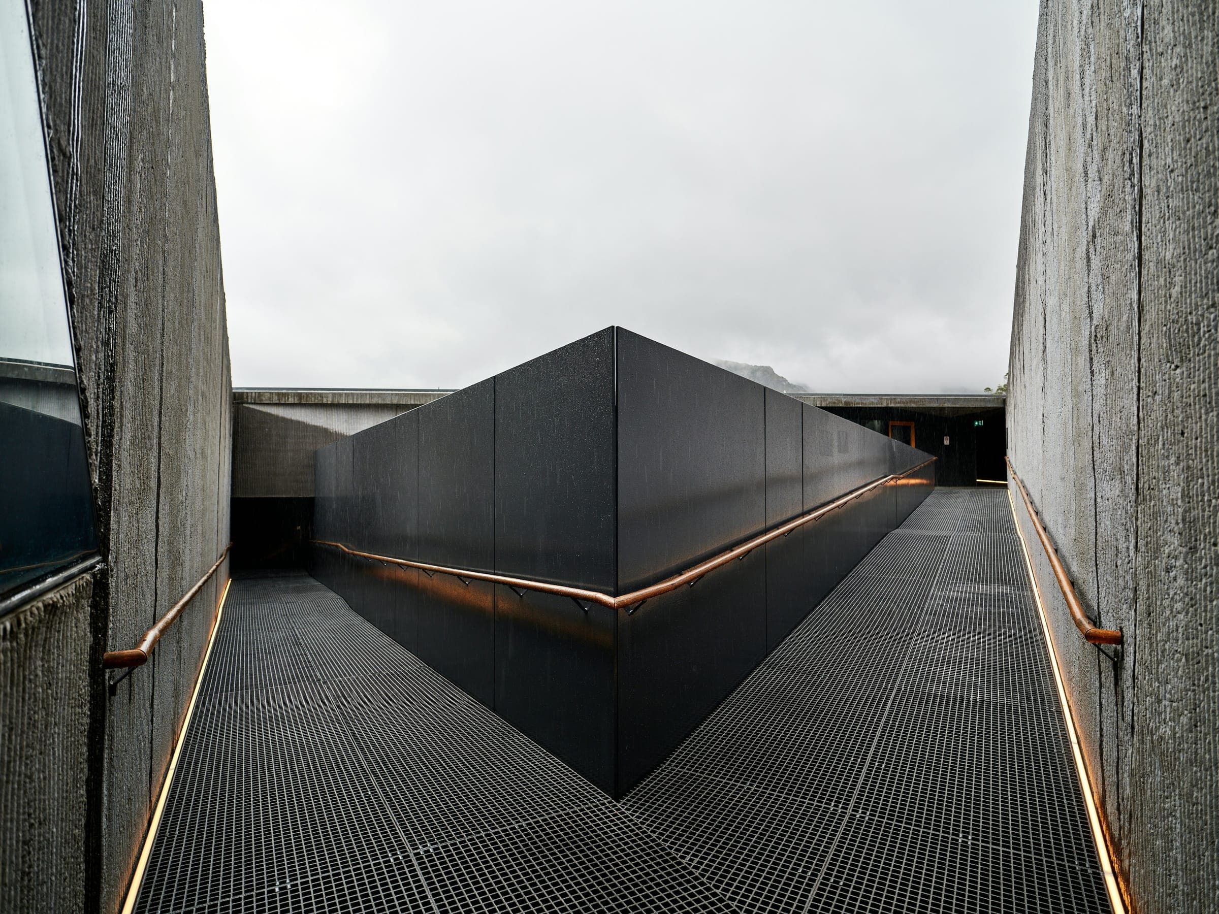 Concrete-walled walkway with a metal grate floor leading around a black angular wall under a grey sky.
