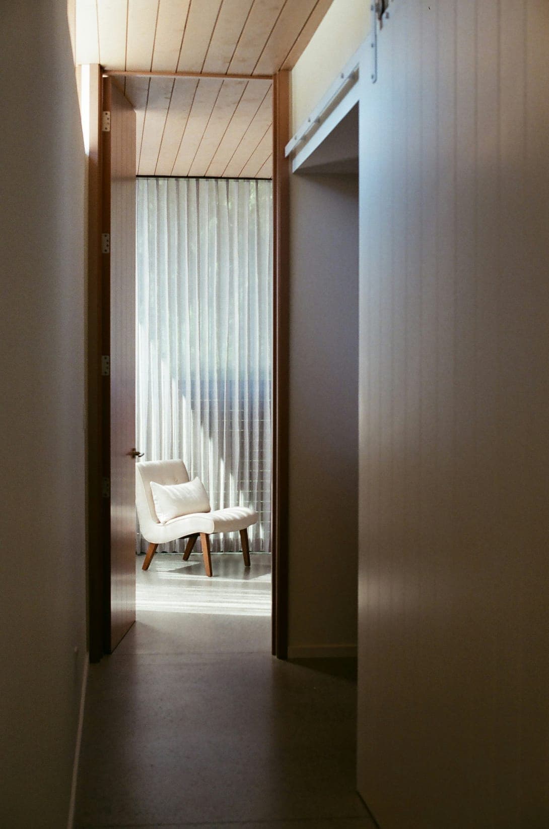 Narrow hallway with a timber ceiling opening onto a room with a white lounge chair before sheer curtains.