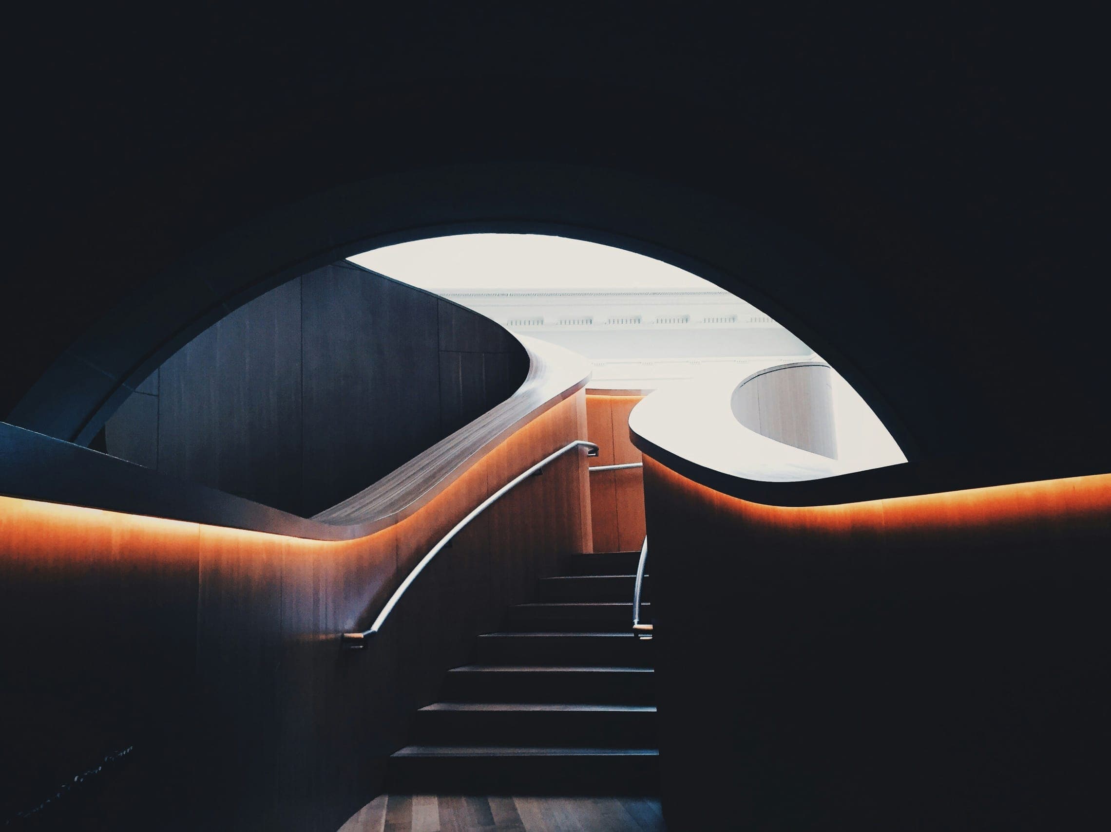 Curving dark staircase with warm hidden lighting rising through an arched opening to daylight.