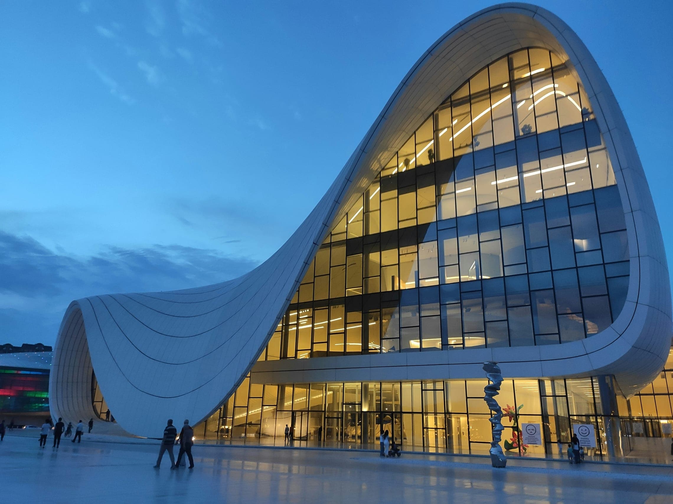 Flowing white building with a vast glass facade lit at dusk, people on the plaza.