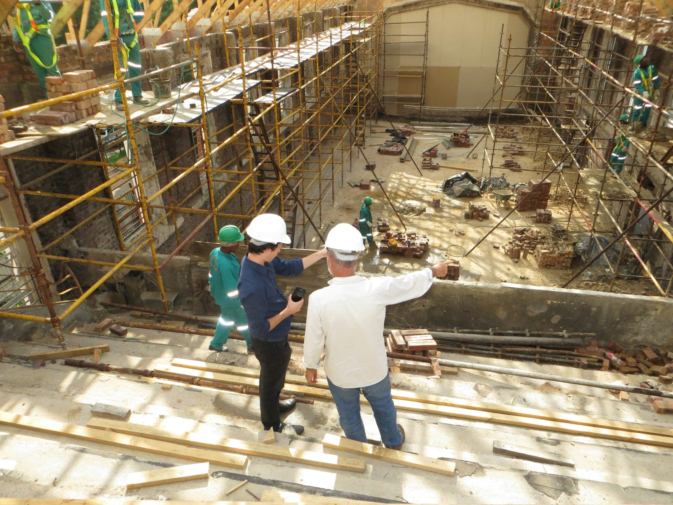 Two people in white hard hats survey a brick building site lined with yellow scaffolding.
