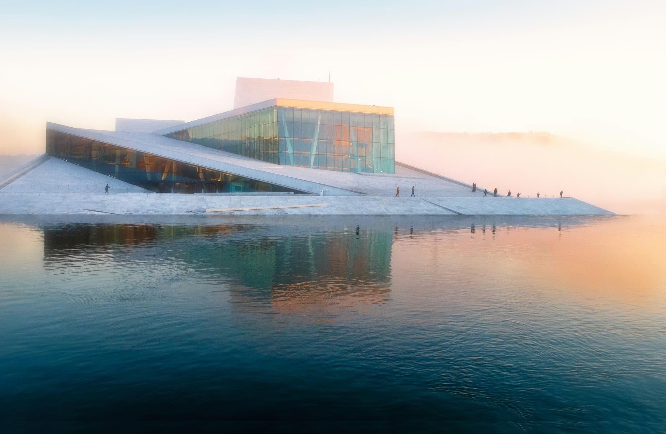 Angular white waterfront building with a glass facade reflected in calm water at misty sunrise.