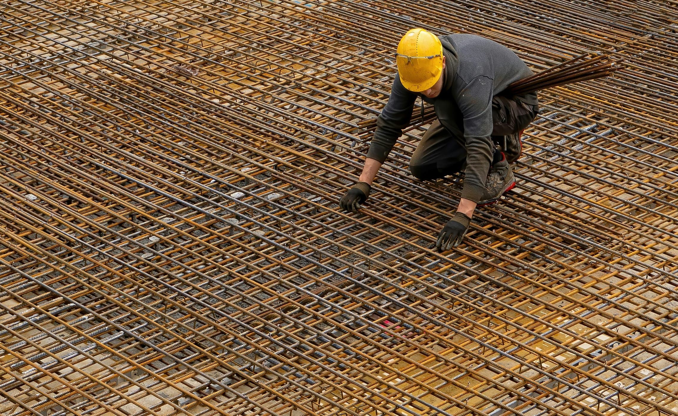 Worker in a yellow hard hat and gloves tying a steel rebar grid.