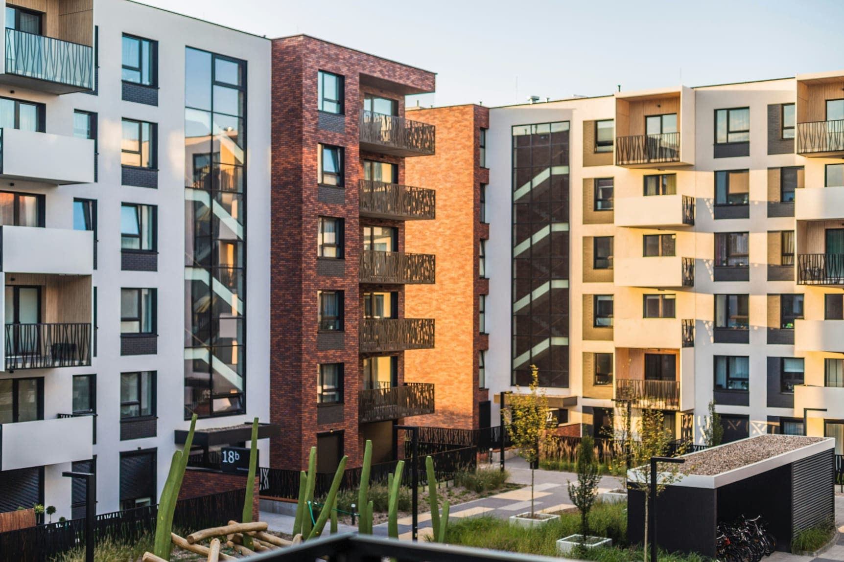 Courtyard framed by white and brick residential blocks with balconies, planting and warm evening light.