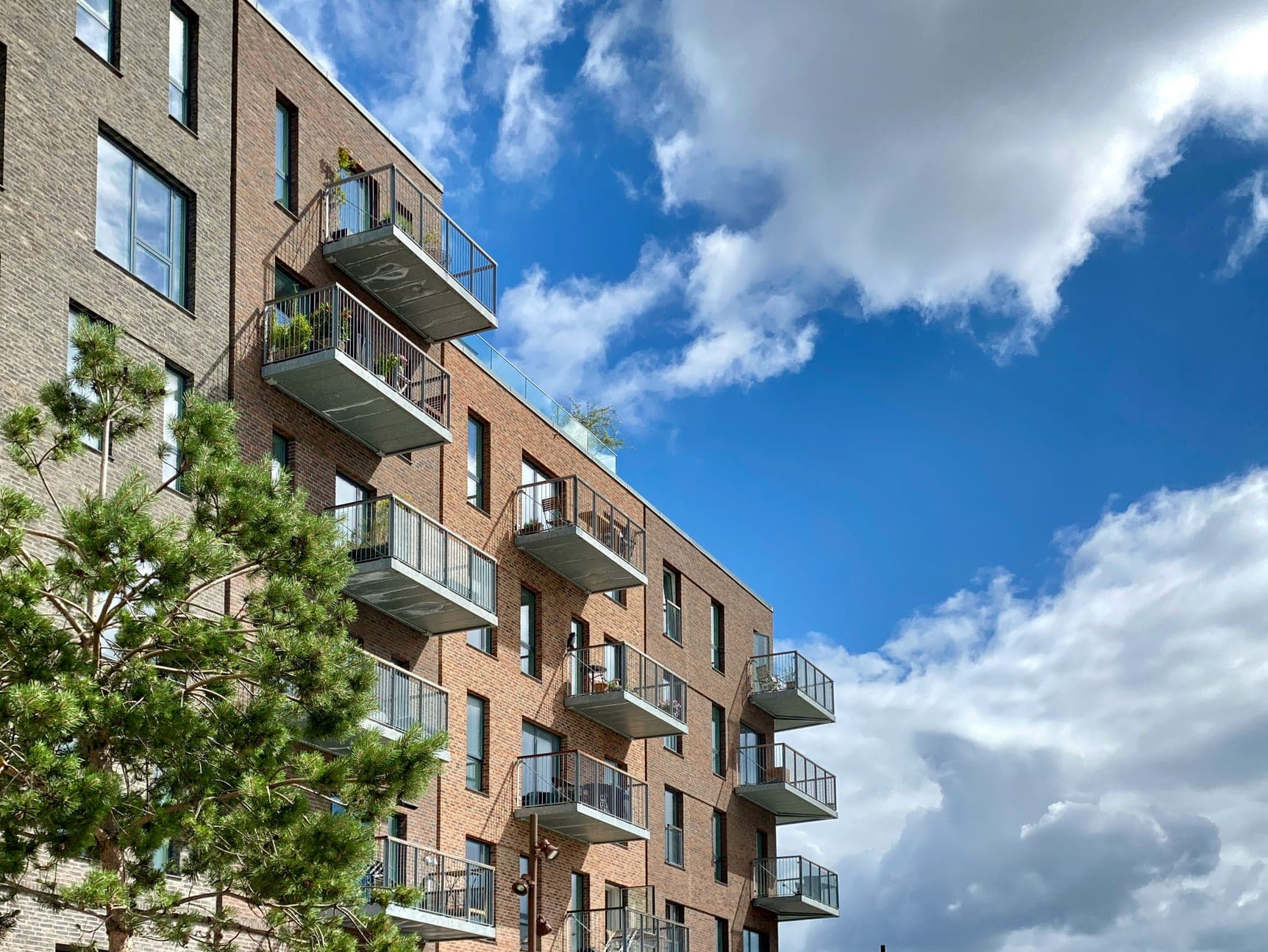 Brick residential facade with projecting glass-railed balconies, a pine tree and bright clouds.
