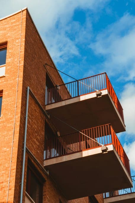 Close view of a brick corner with concrete balconies and rust-red railings under blue sky.