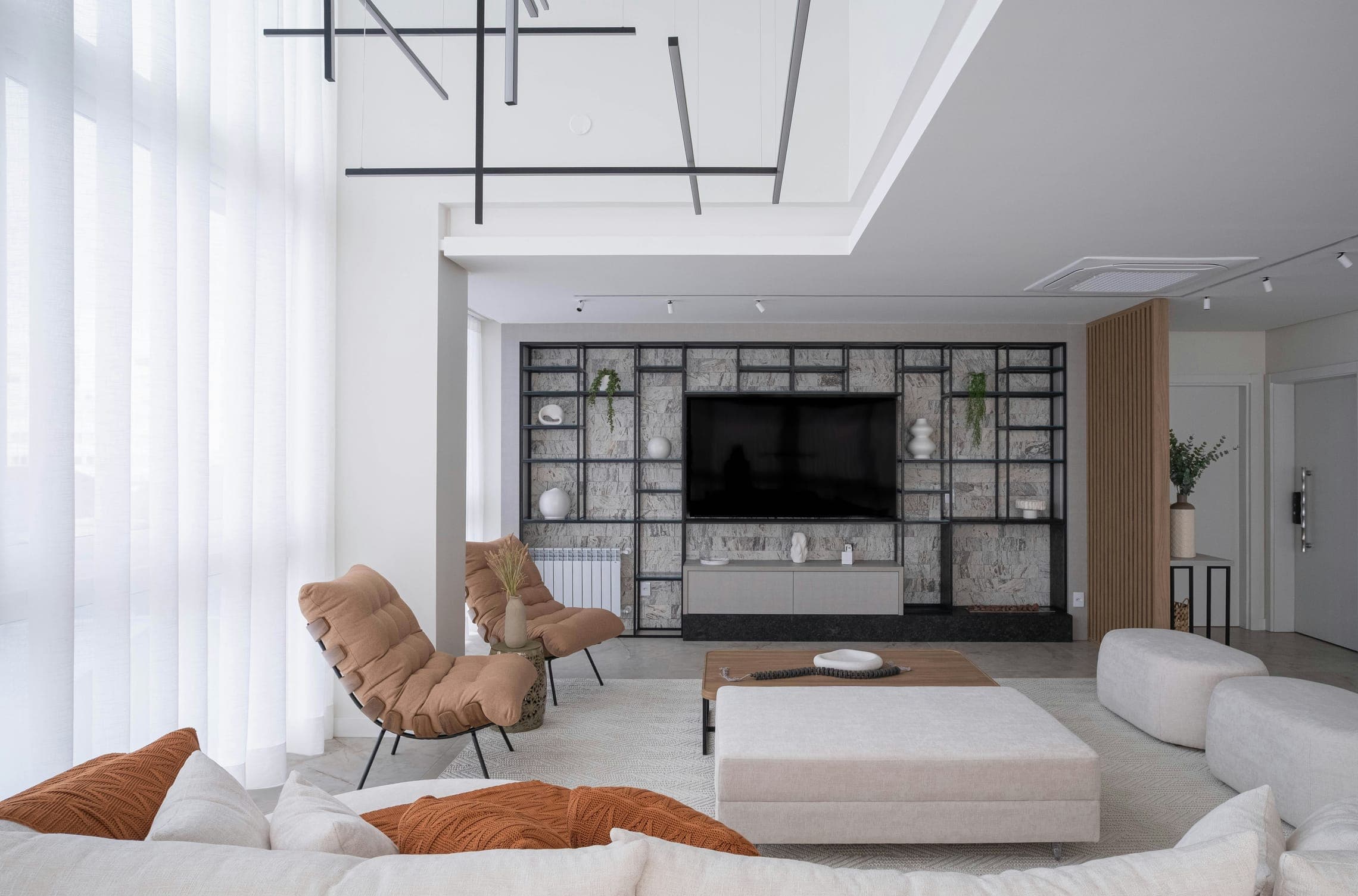 Double-height living room with sheer curtains, rust lounge chairs, white ottoman and shelving wall.