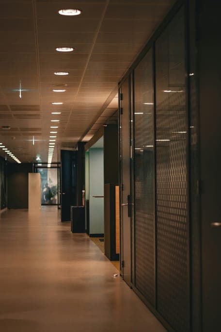 Dim office corridor lined with glass partitions and rows of recessed ceiling lights.