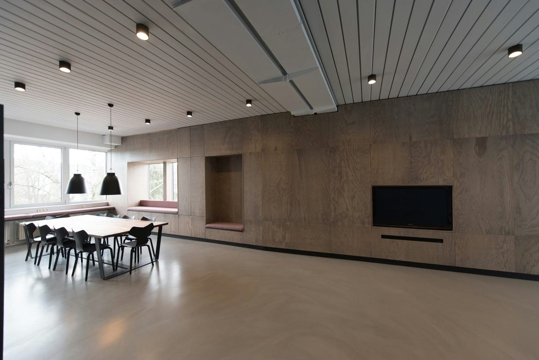 Workspace with plywood-clad walls, long dining table, black chairs and pendant lights.