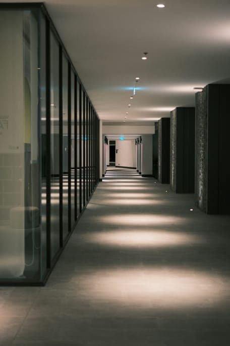Long dark corridor with glass walls on one side and pools of light on the floor.