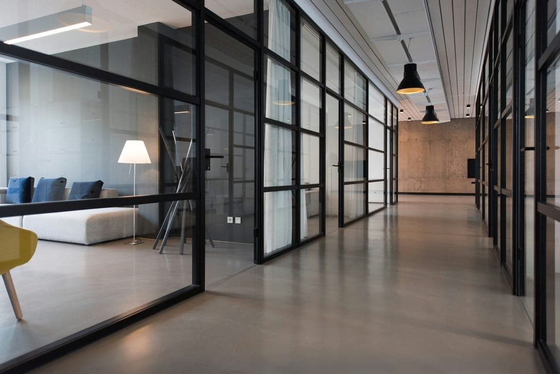 Corridor of black-framed glass offices on polished concrete, a lounge visible through the glass.