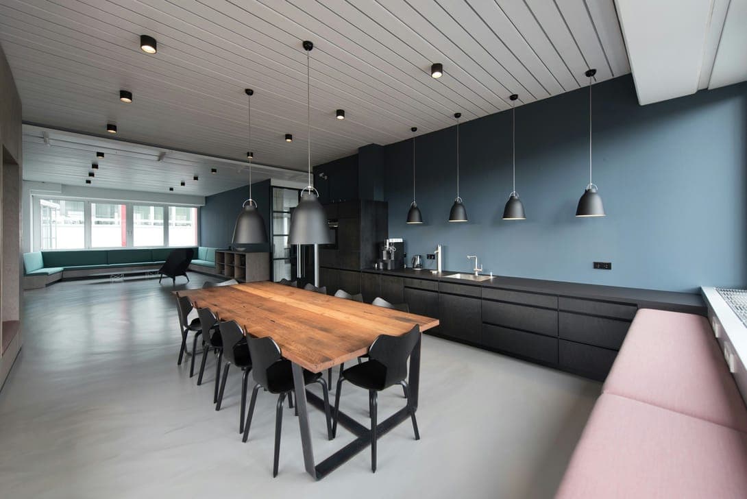 Office kitchen with blue wall, black cabinets, wooden table, black pendants and pink bench.
