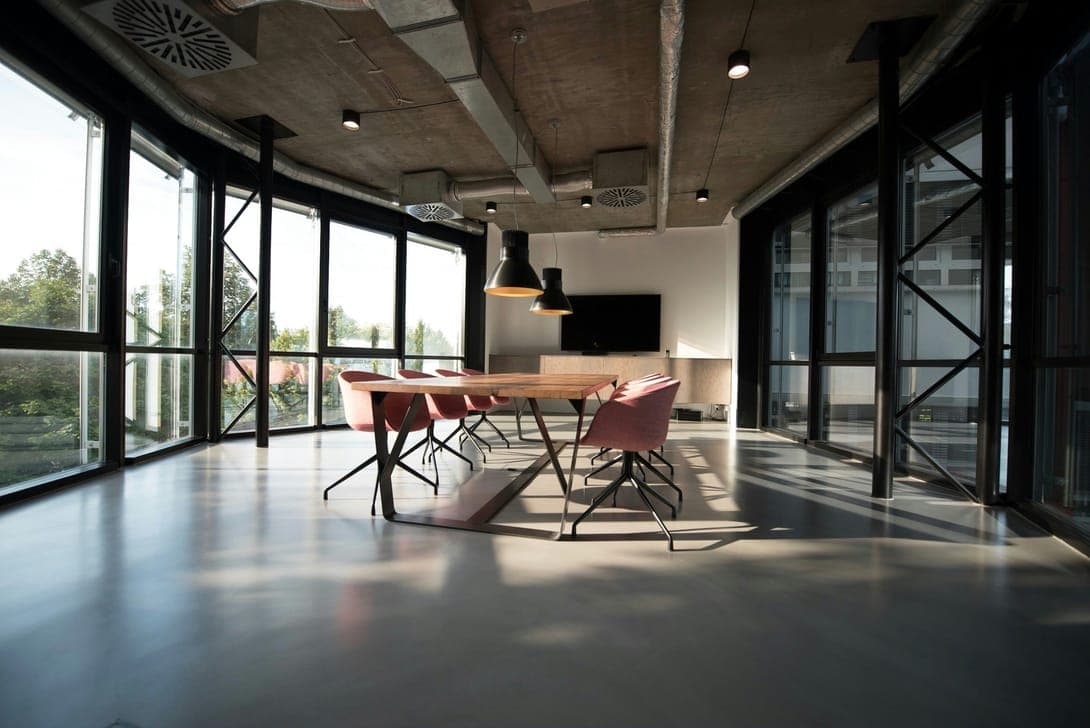 Sunlit meeting room with wooden table, pink chairs, exposed concrete ceiling and tall windows.