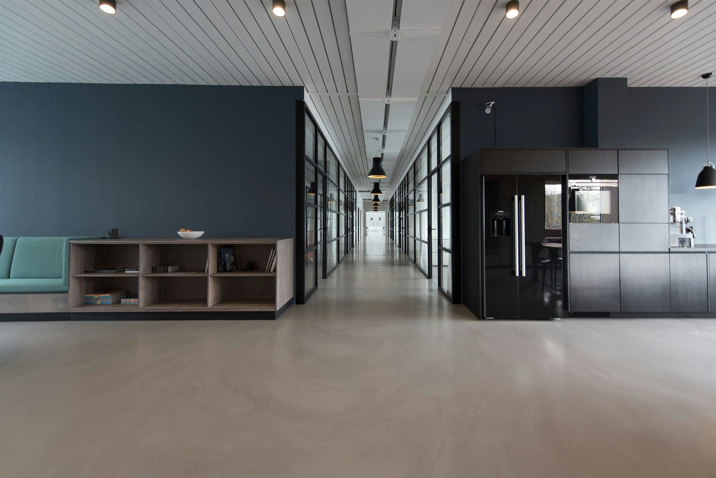 Open office floor with concrete flooring, dark kitchen cabinetry and a long glazed corridor.