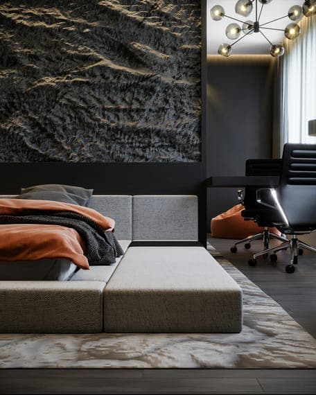 Dark bedroom with low grey platform bed, orange bedding, textured wall panel and desk.