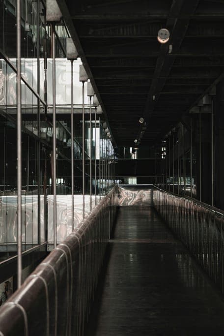 Narrow walkway with curved railings beside glass walls under a dark steel roof.
