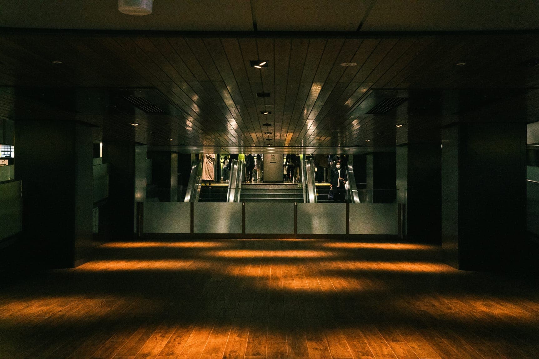 Dim concourse with timber floor striped by sunlight, escalators and people in the distance.