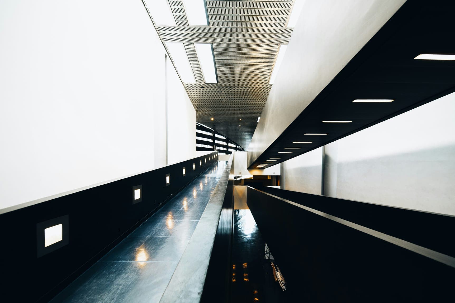 Long white interior ramp with dark glossy floor, small wall lights and skylit ceiling.