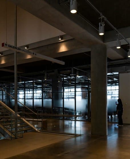 Concrete hall with track lights, glass rooms, open staircase and a hanging directional sign.