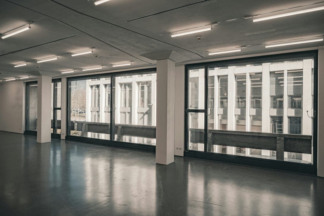Empty open floor with tall windows, white columns and fluorescent strip lights.