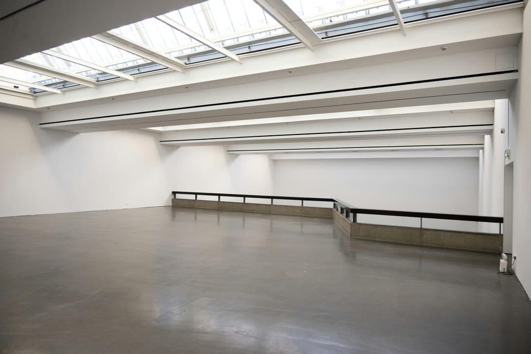 Empty white gallery under a large skylight, grey concrete floor and a low railing.