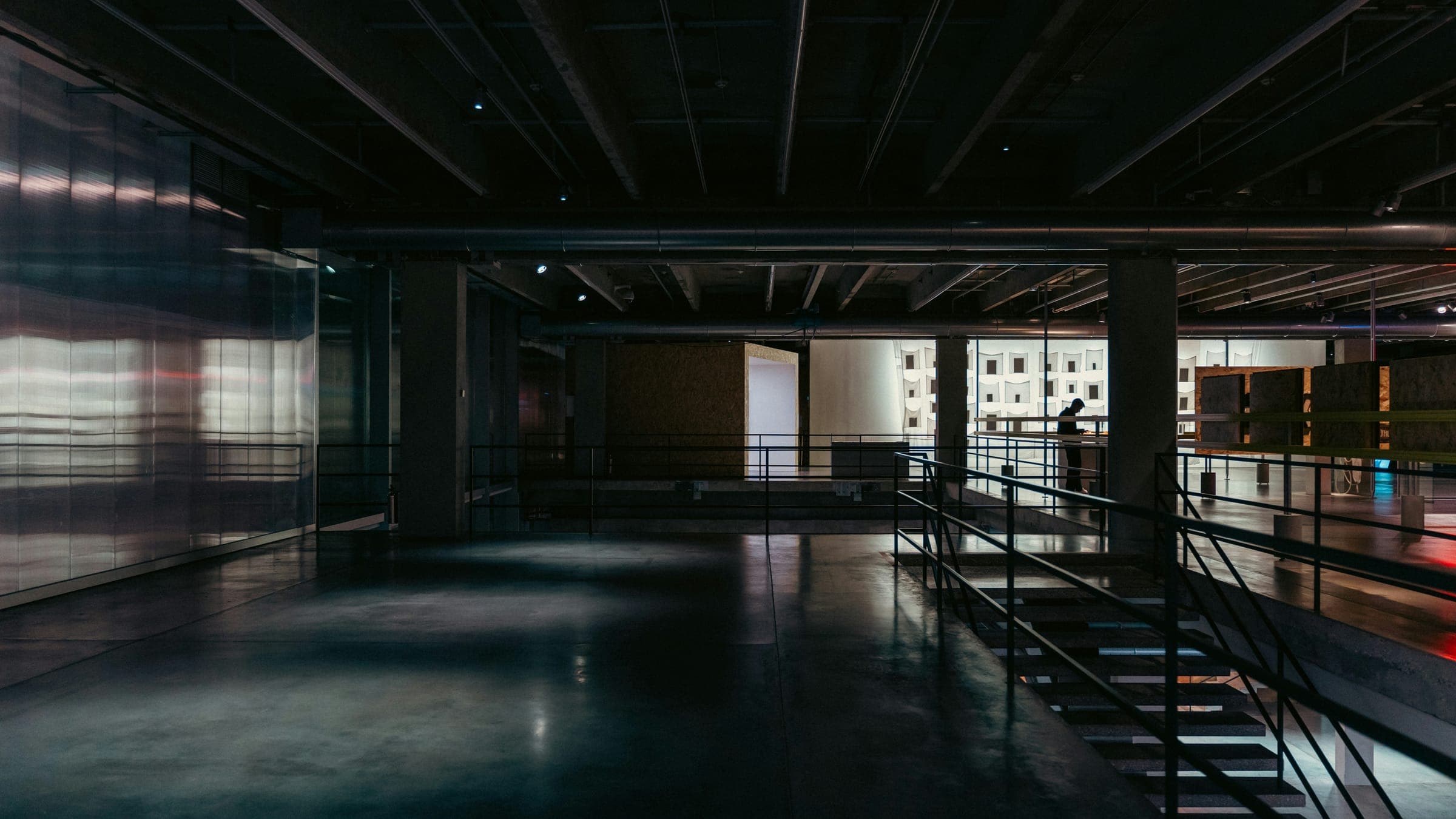 Dark open hall with exposed beams, glossy concrete floor, steel staircase and a distant figure.