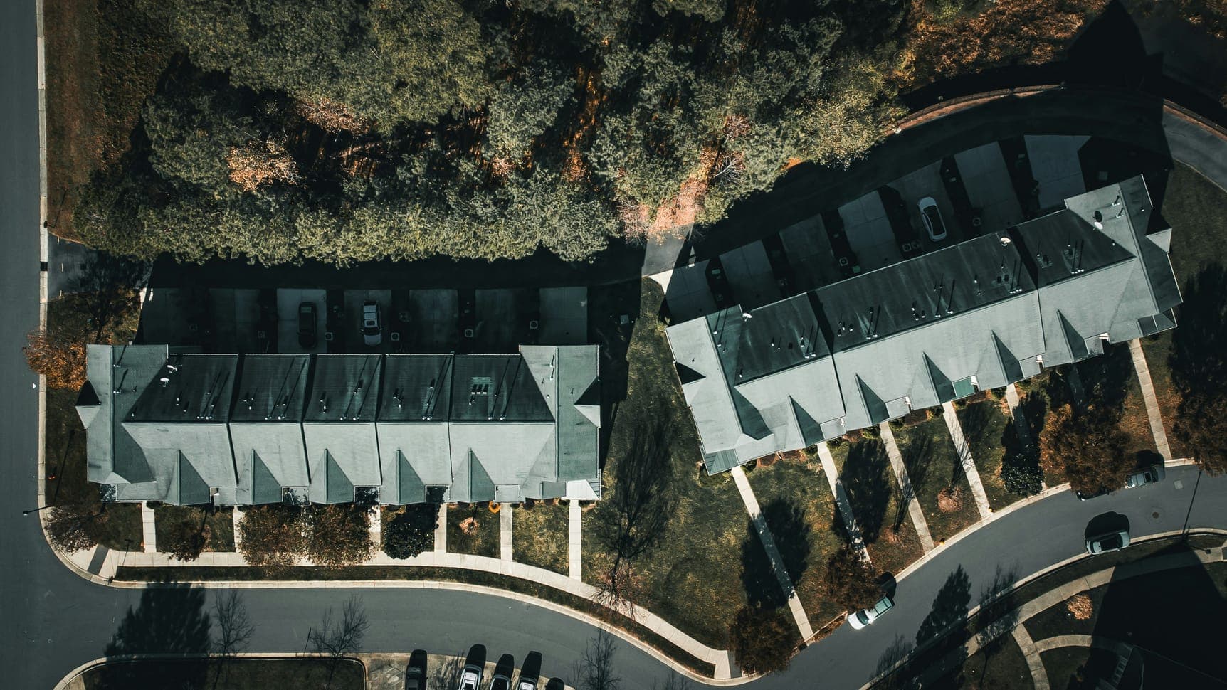 Aerial view of two rows of grey-roofed townhouses beside trees and a curving road.