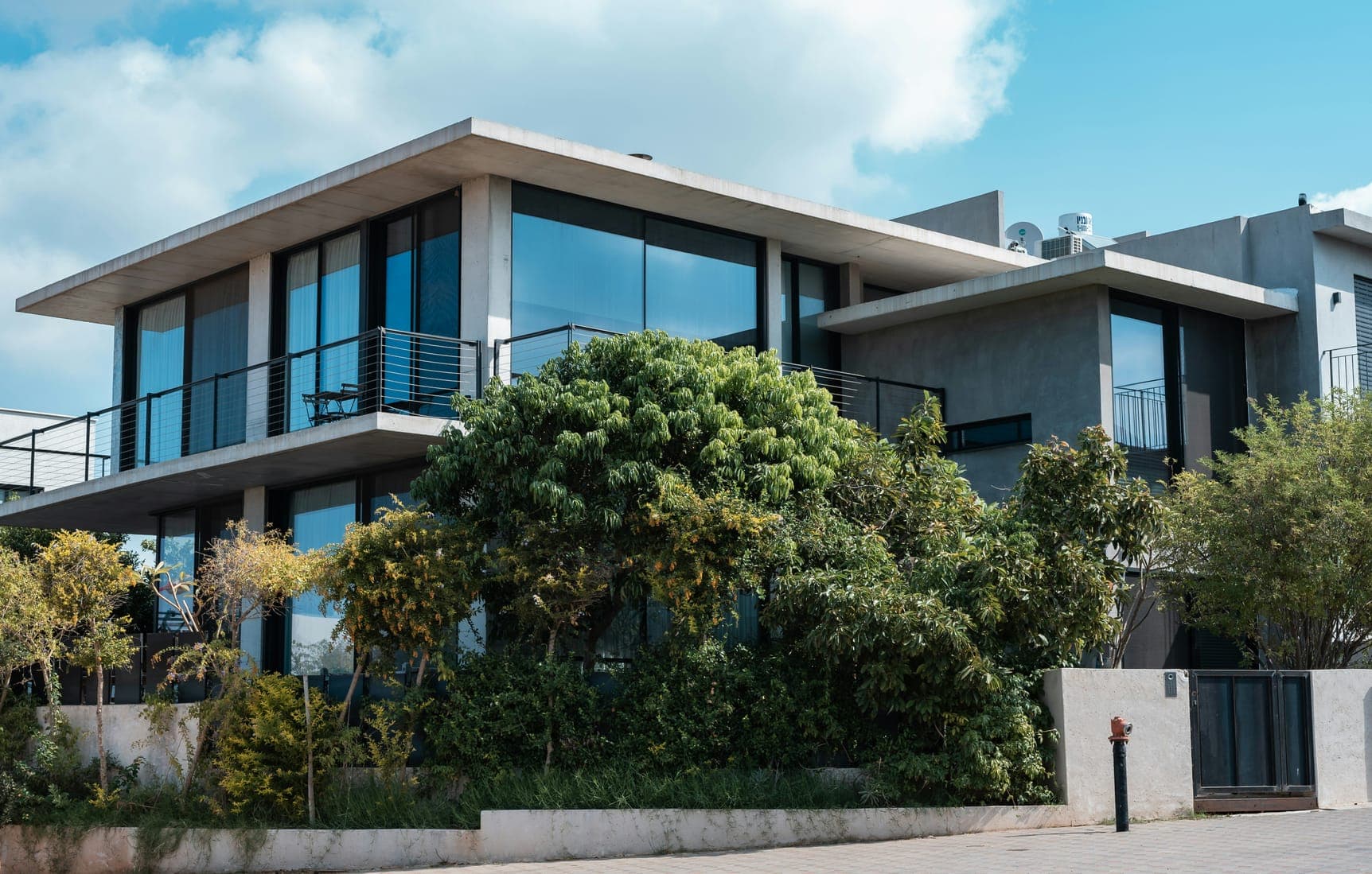 Modern two-storey concrete house with large dark windows, balconies and lush front garden.