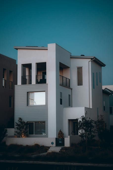 White three-storey modern house with balconies and lit windows against a teal dusk sky.