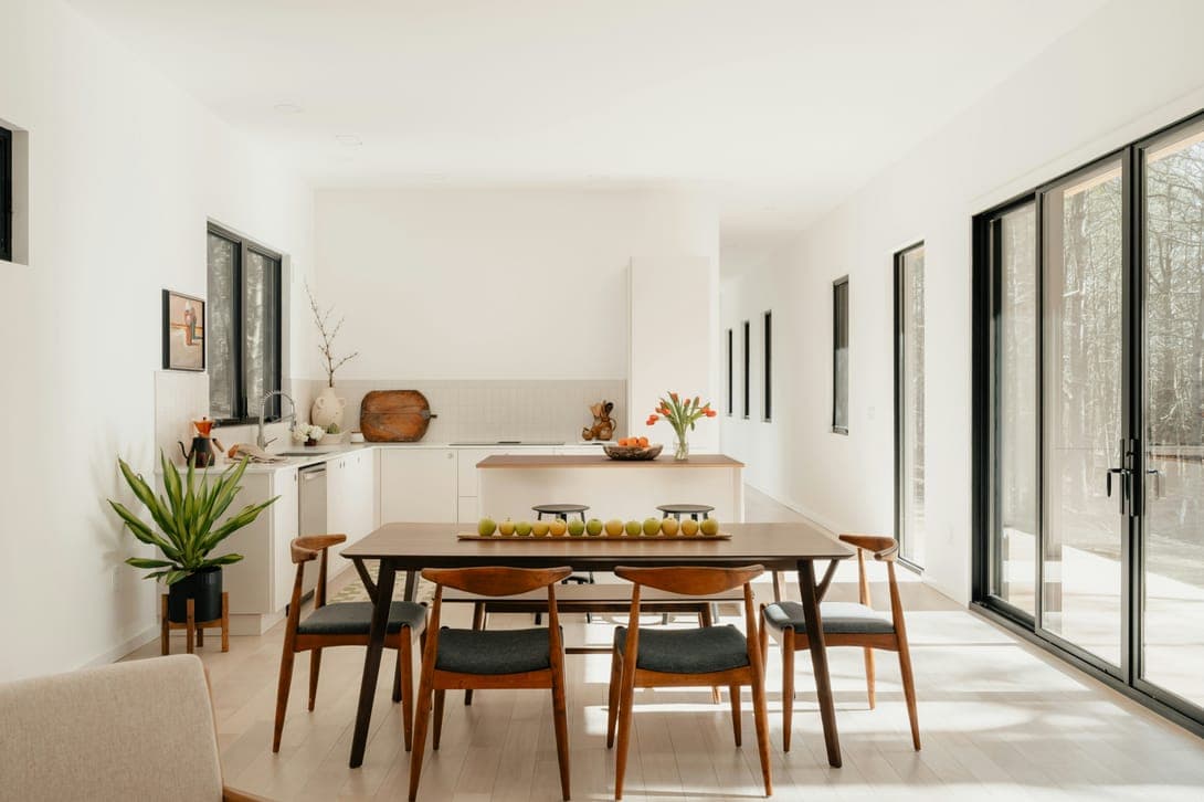 Sunlit open kitchen and dining room with wooden table, mid-century chairs and glass doors.