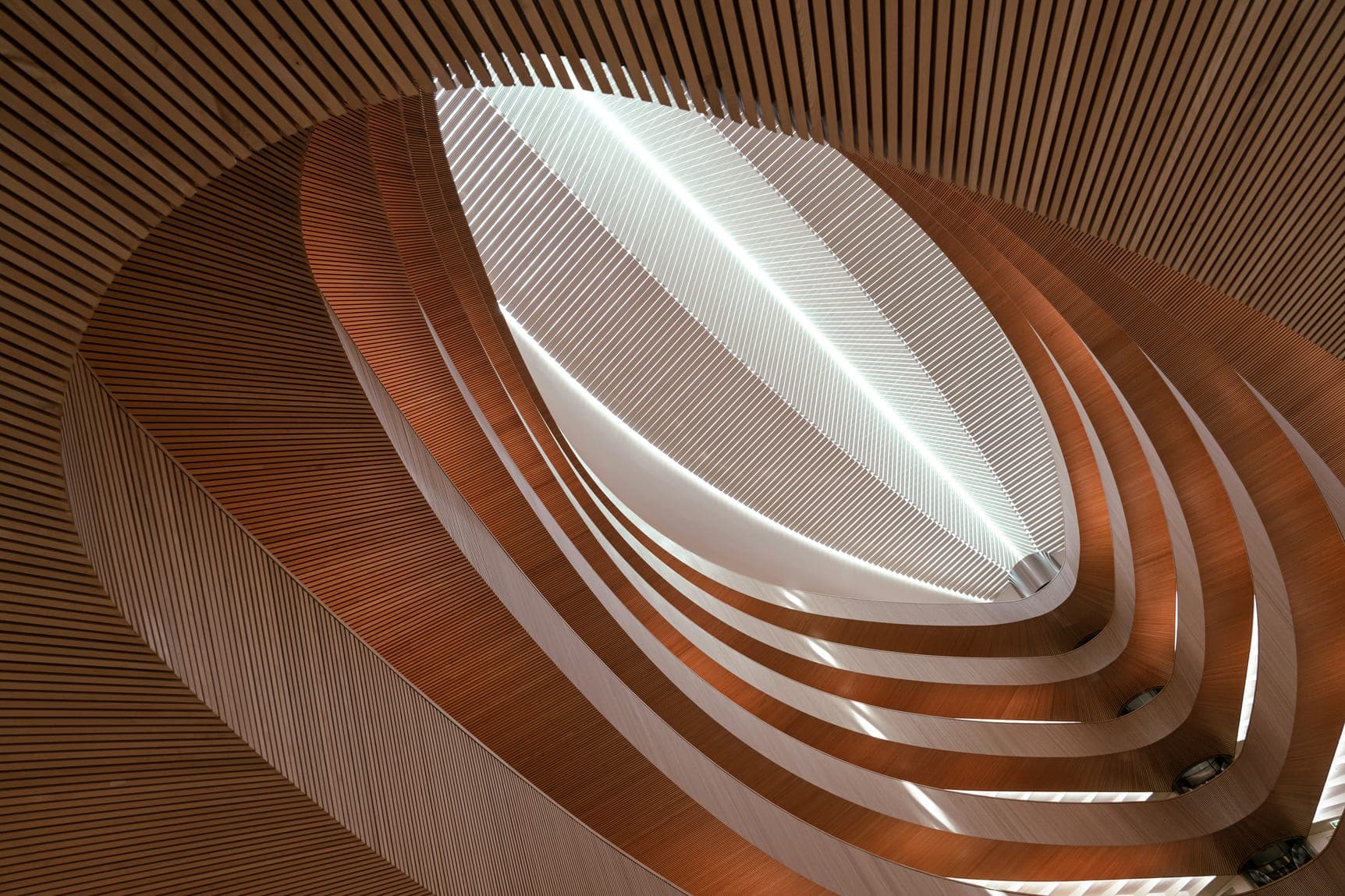 Upward view of layered wooden balconies wrapping an elliptical skylight in soft light.