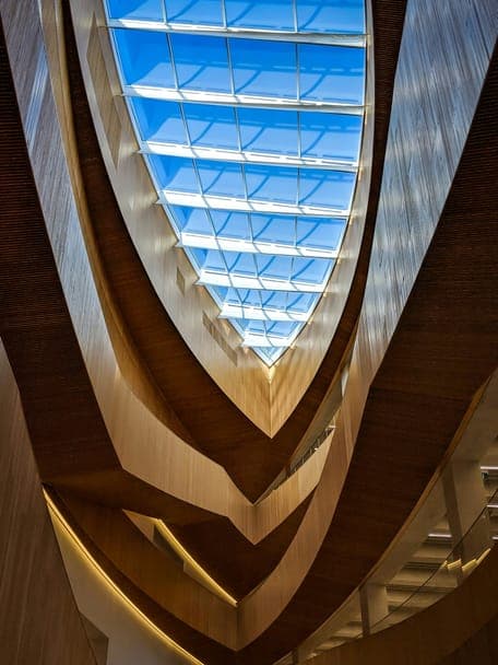Upward view through curved wooden atrium levels to a blue-lit glass roof.