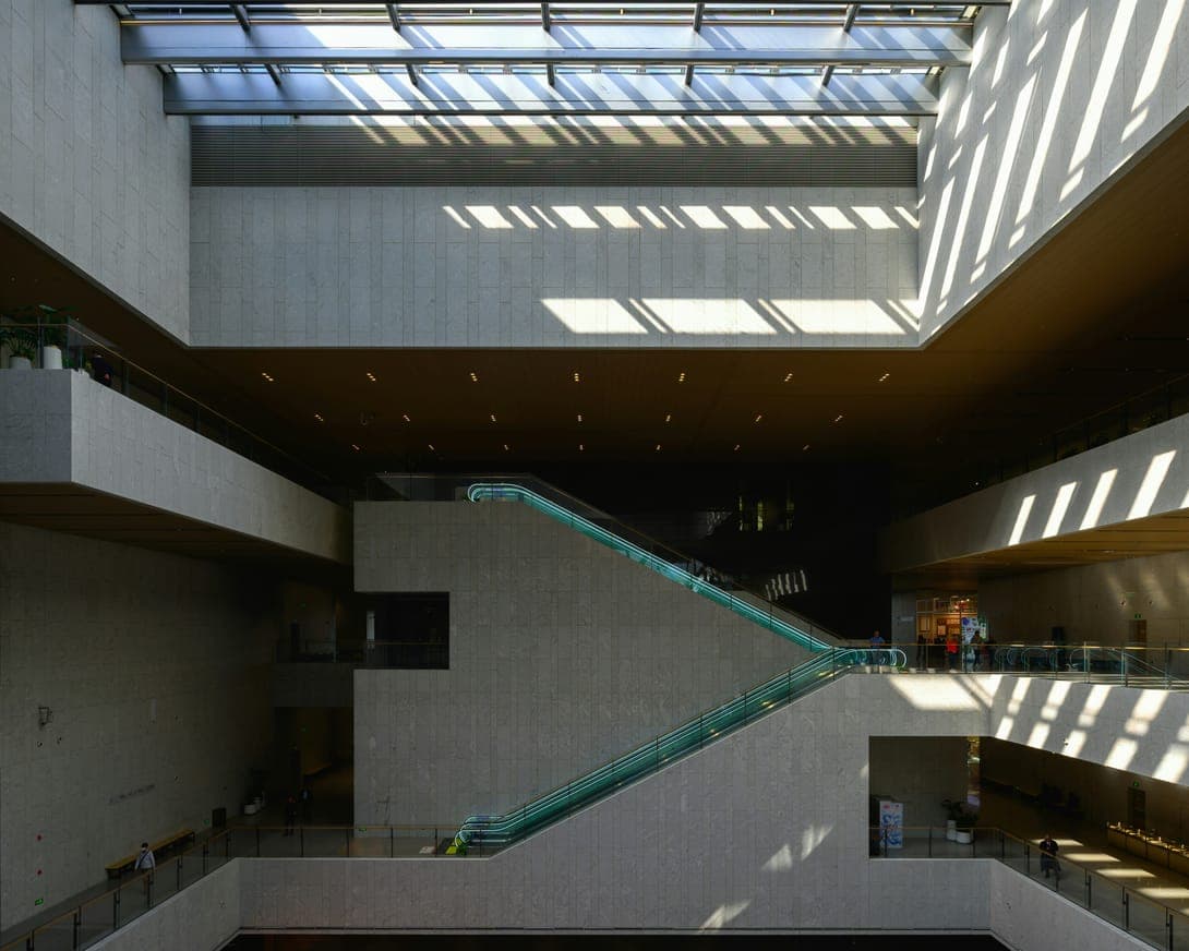 Stone-clad atrium with skylight shadows, criss-crossing escalators and balconies on several levels.