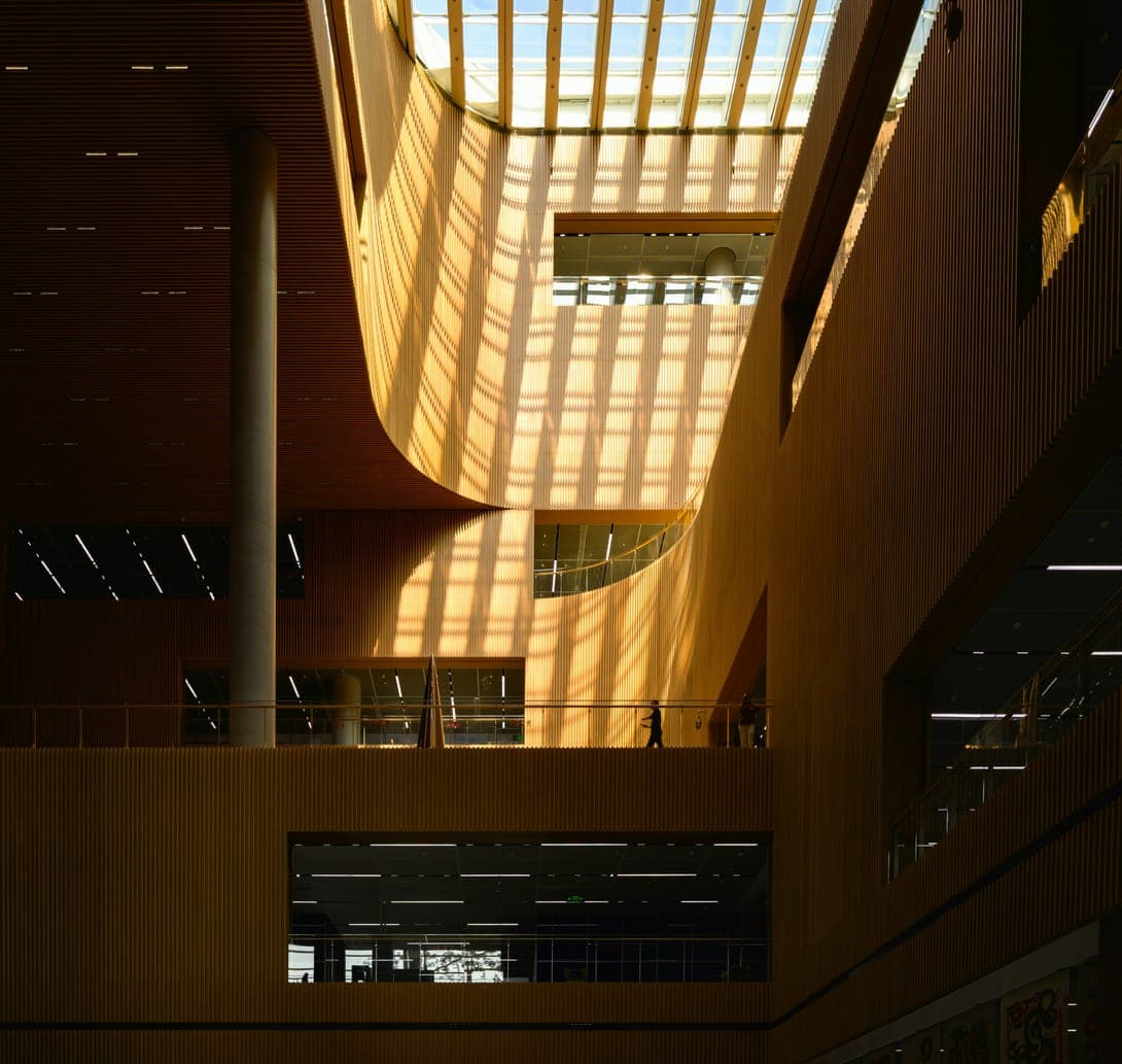 Tall atrium of timber slats lit by skylight stripes, people on an upper walkway.
