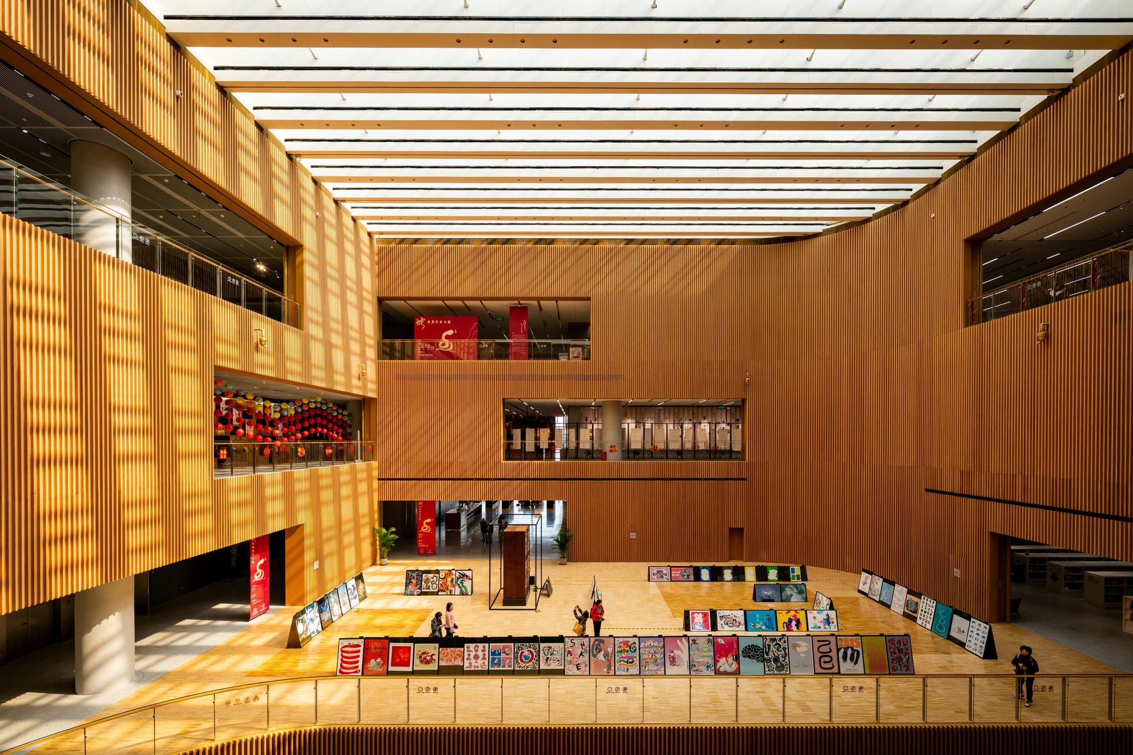 Timber-lined atrium with a colourful poster exhibition on the floor and red banners above.