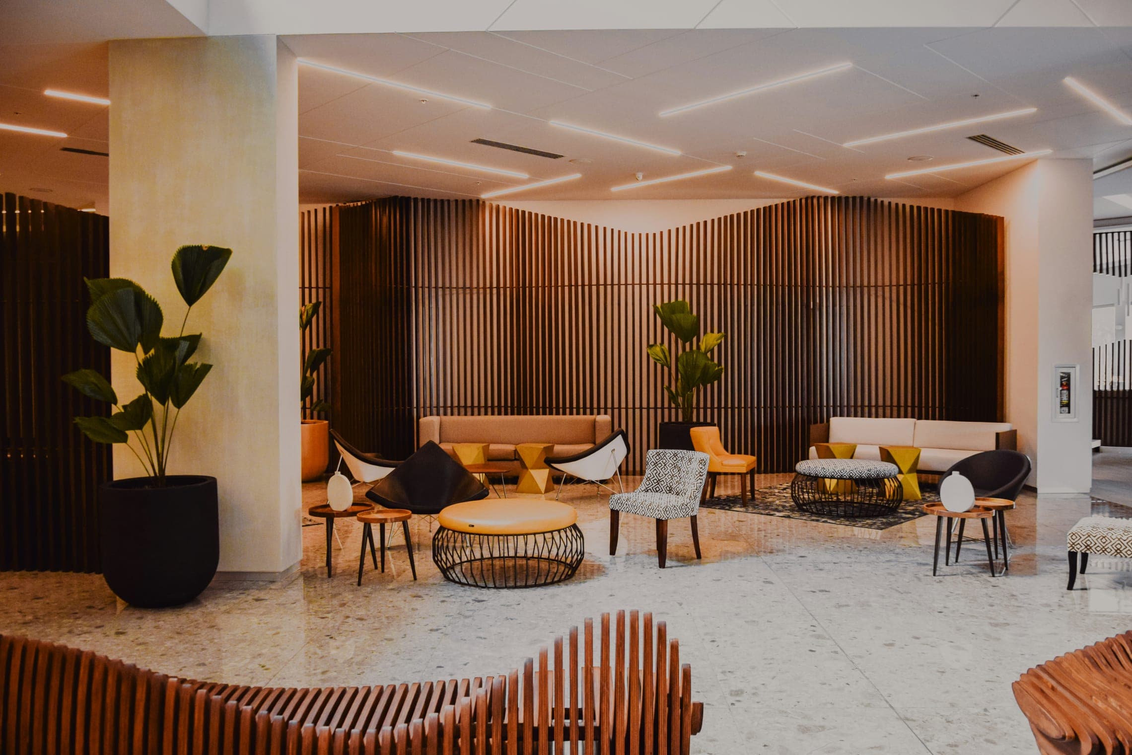 Lobby lounge with slatted timber wall, terrazzo floor, mixed seating and potted plants.