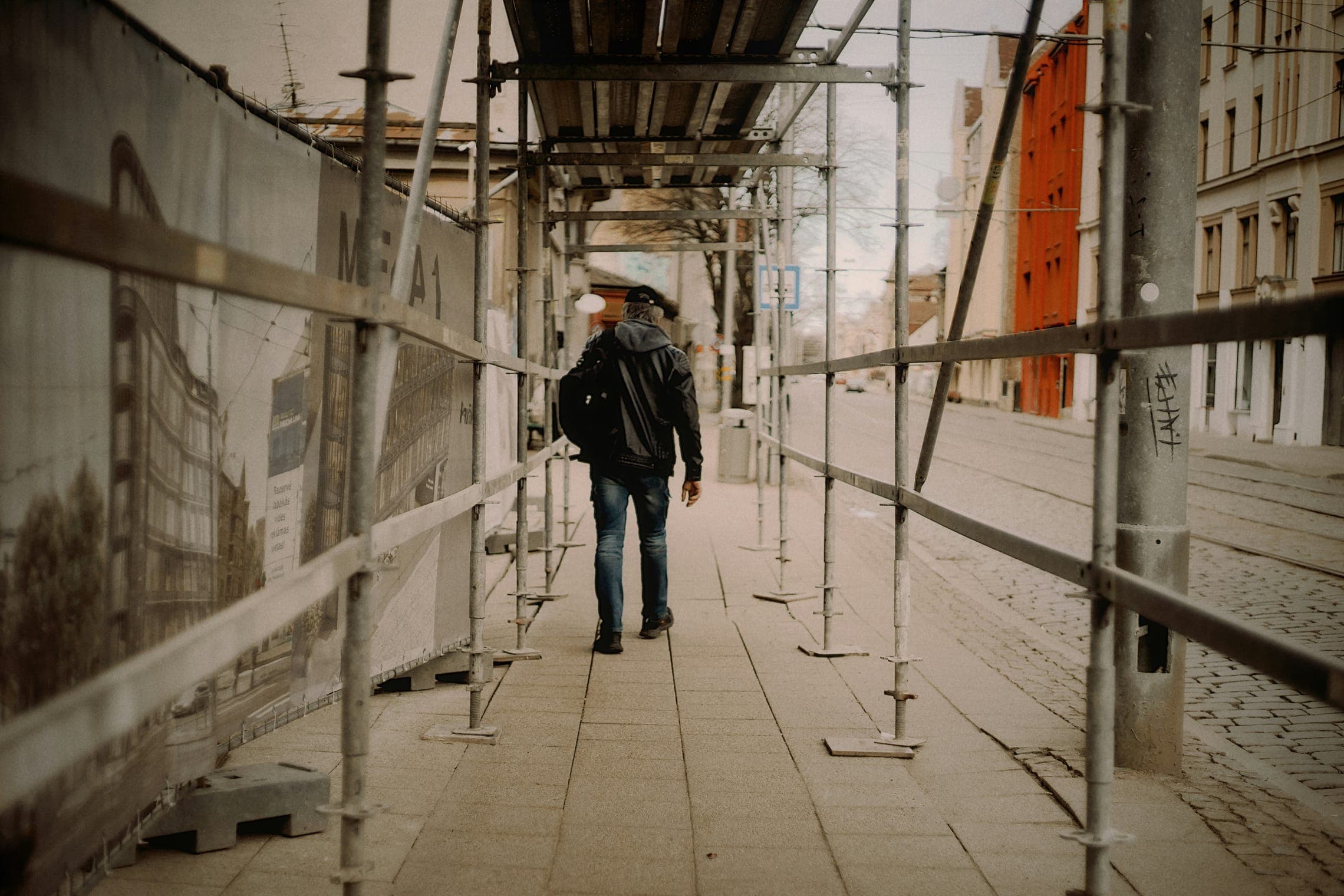 Person walking under scaffolding along a covered sidewalk beside printed construction hoarding.