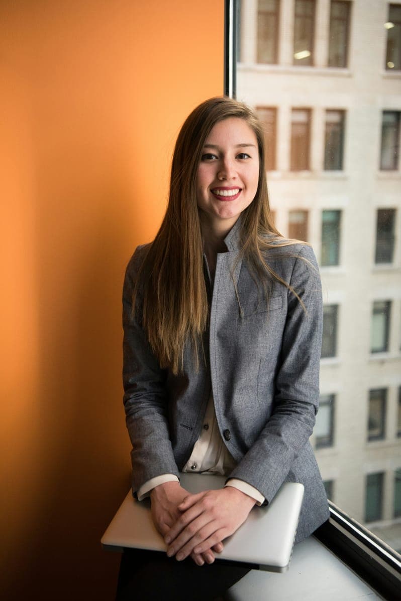Portrait of Ava Mitchell, smiling woman in a grey blazer holding a laptop by a window.