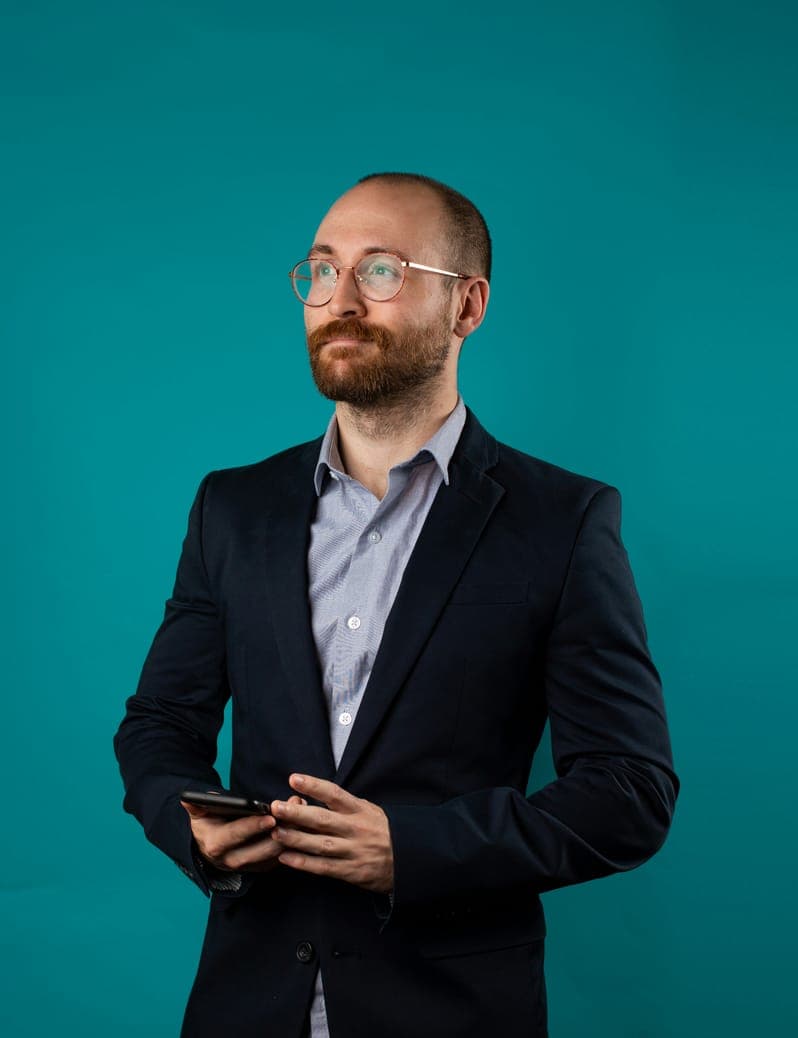 Portrait of Ethan Carter, bearded man in glasses and navy blazer holding a phone, teal backdrop.