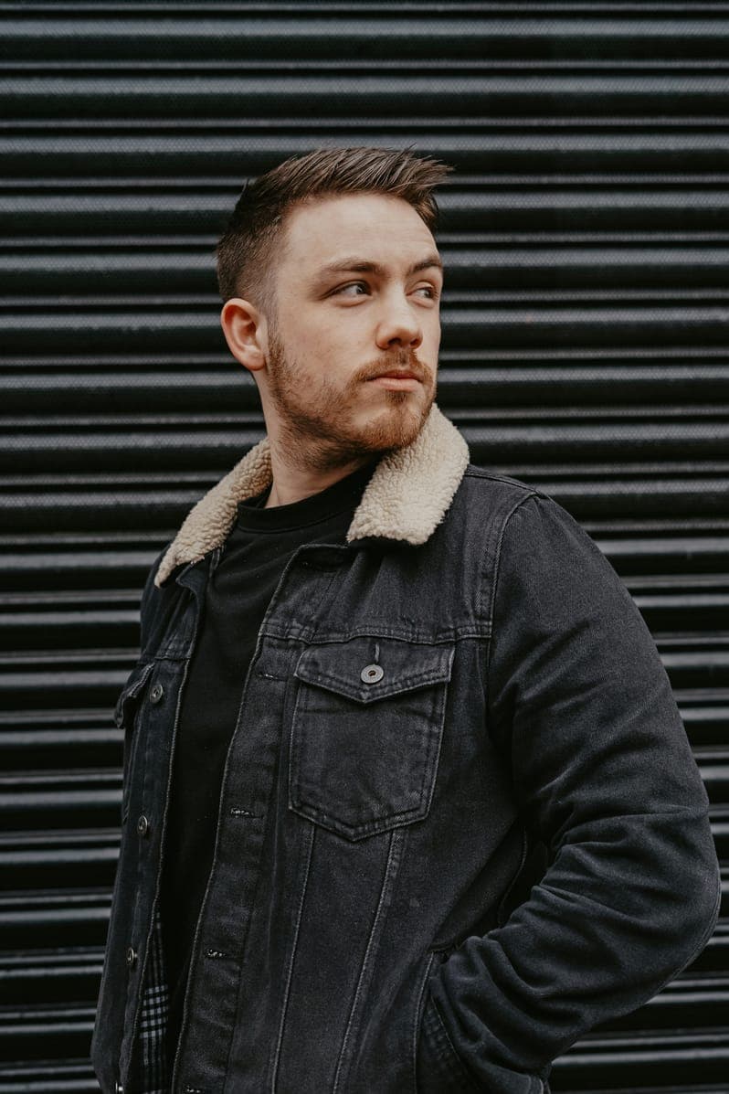 Portrait of Noah Bennett, bearded man in a sherpa-collar denim jacket before a metal shutter.
