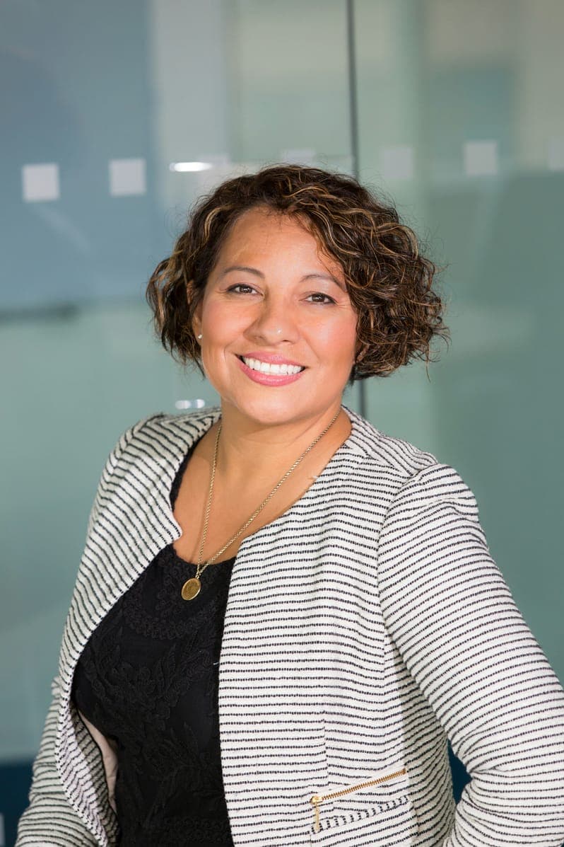 Portrait of Olivia Turner, smiling woman with curly hair in a striped jacket by glass.